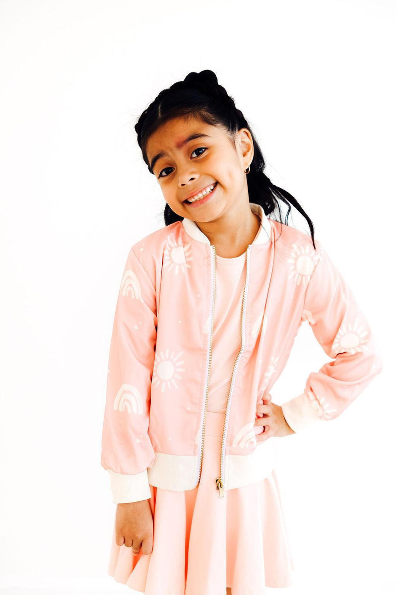 A smiling girl wearing the Soft Pink Sun Satin Jacket by Mila & Rose, posed confidently with her hands on her hips. The jacket features cheerful sun and rainbow designs, perfect for stylish little girls.