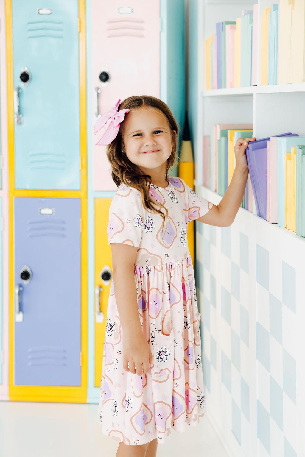Happy child in a Peanut Butter Jelly Time S/S Pocket Twirl Dress by Mila & Rose, standing next to colorful lockers, smiling while reaching for a book.