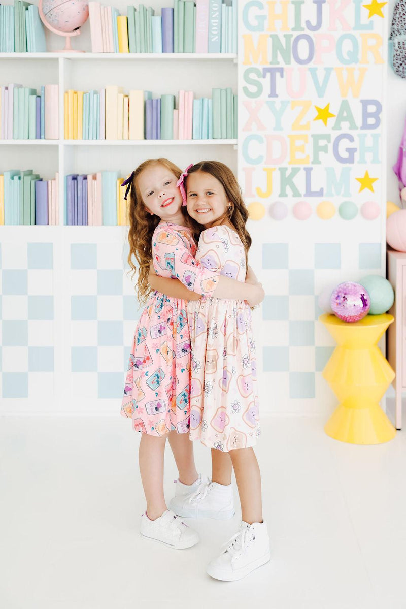 Two joyful girls in the Peanut Butter Jelly Time S/S Pocket Twirl Dress by Mila & Rose, happily hugging each other against a playful background filled with colorful books.
