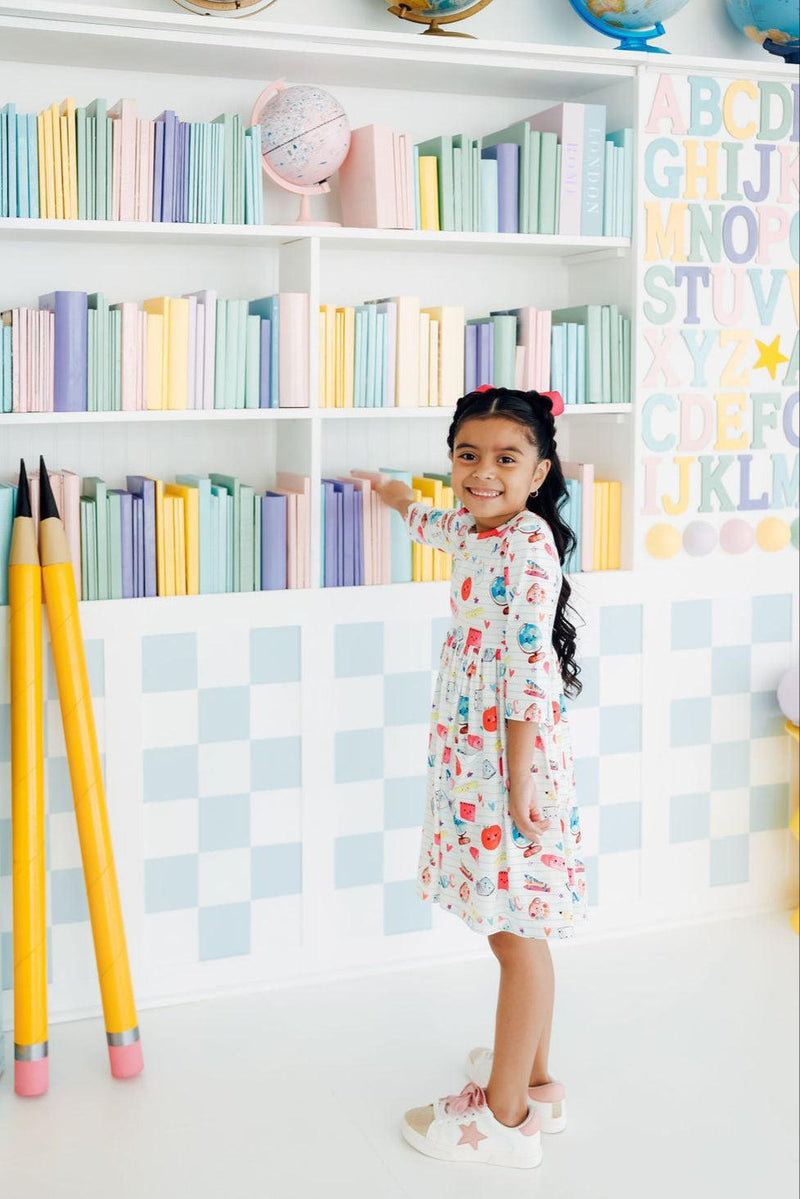 Little girl wearing the Smarty Pants Pocket Twirl Dress, playfully standing in front of a colorful bookshelf filled with books.