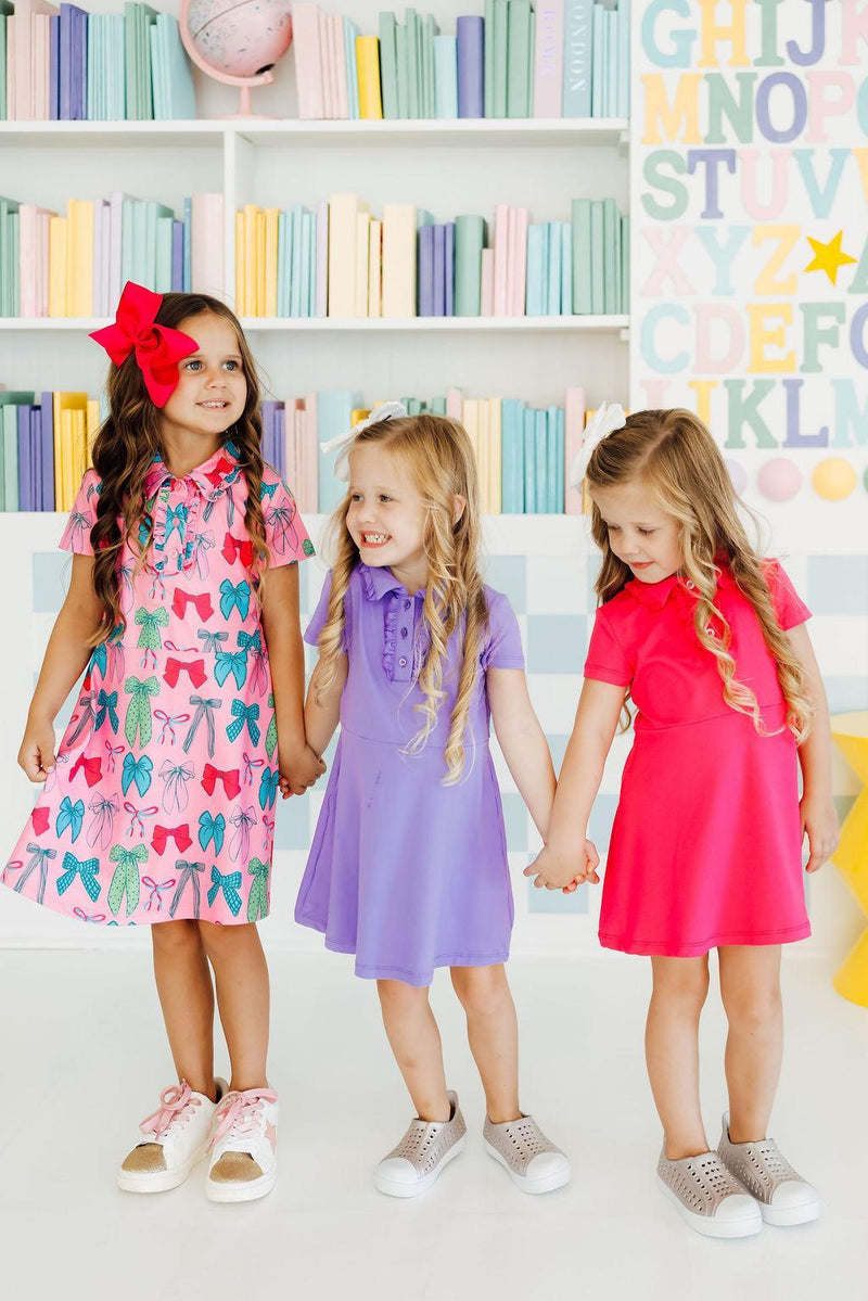 Three adorable girls in vibrant dresses, including the Periwinkle S/S Polo Dress by Mila & Rose®, holding hands in a colorful setting—perfect for playdates and fun times!