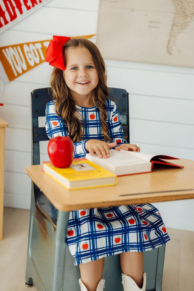 A girl sitting at a desk in the Plaid Apples L/S Pocket Twirl Dress, joyfully reading a book with an apple and colorful books nearby.
