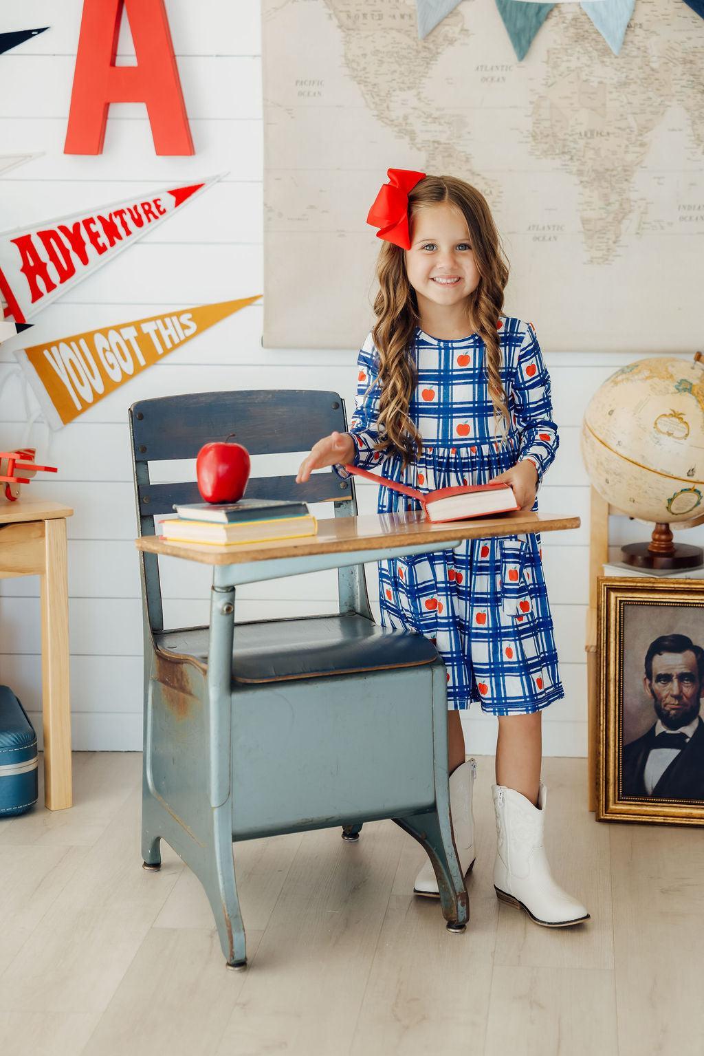 Plaid Apples L/S Pocket Twirl Dress worn by a smiling girl in a classroom setting, complete with an apple and books, embodying playful learning.
