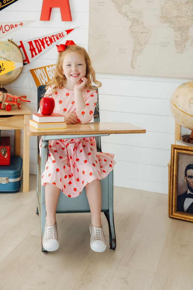 A smiling girl in an 'Apple a Day' 3/4 Sleeve Pocket Girls Twirl Dress, sitting at a desk with an apple and books, capturing a delightful school day look.
