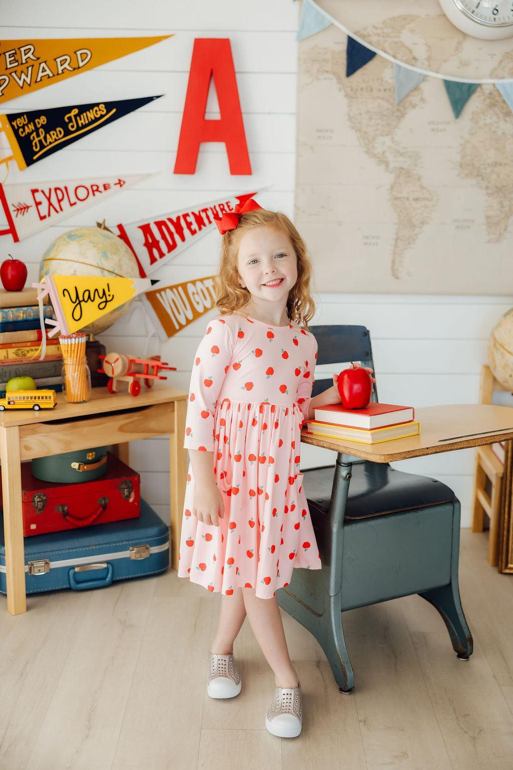 A cheerful girl wearing an 'Apple a Day' 3/4 Sleeve Pocket Girls Twirl Dress, standing beside a desk with an apple and books, showcasing a fun and playful style.