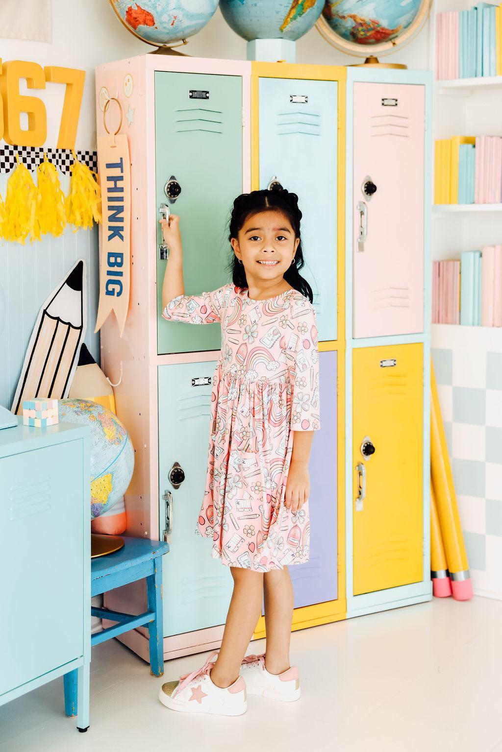 A smiling girl wearing the Dream Big Little One 3/4 Pocket Twirl Dress by Mila & Rose® standing next to colorful lockers, showcasing the playful and vibrant design of the dress.