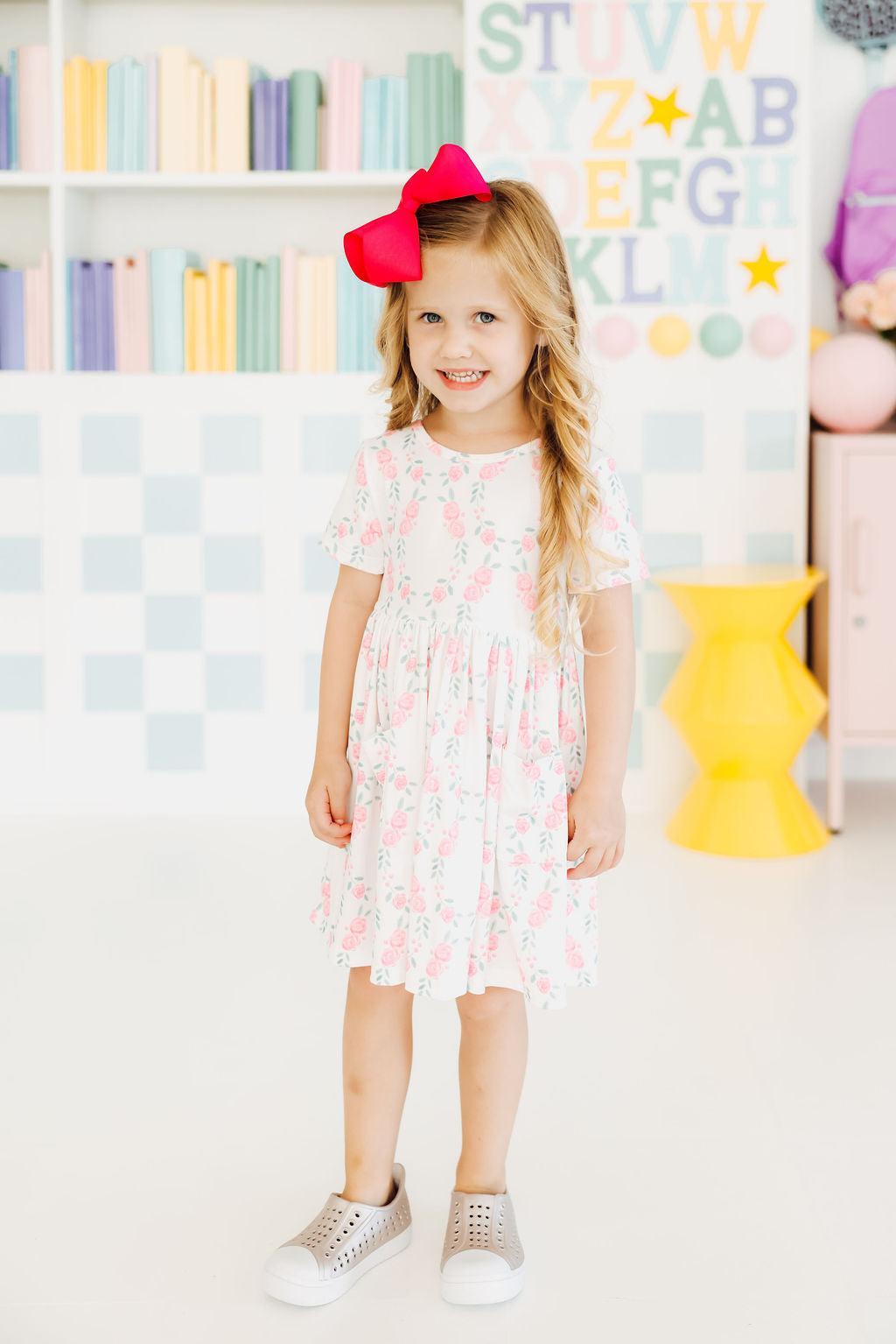 A cheerful little girl wearing the Rosie Posie S/S Pocket Twirl Dress by Mila & Rose, twirling happily in a garden, showcasing its playful design and vibrant colors.