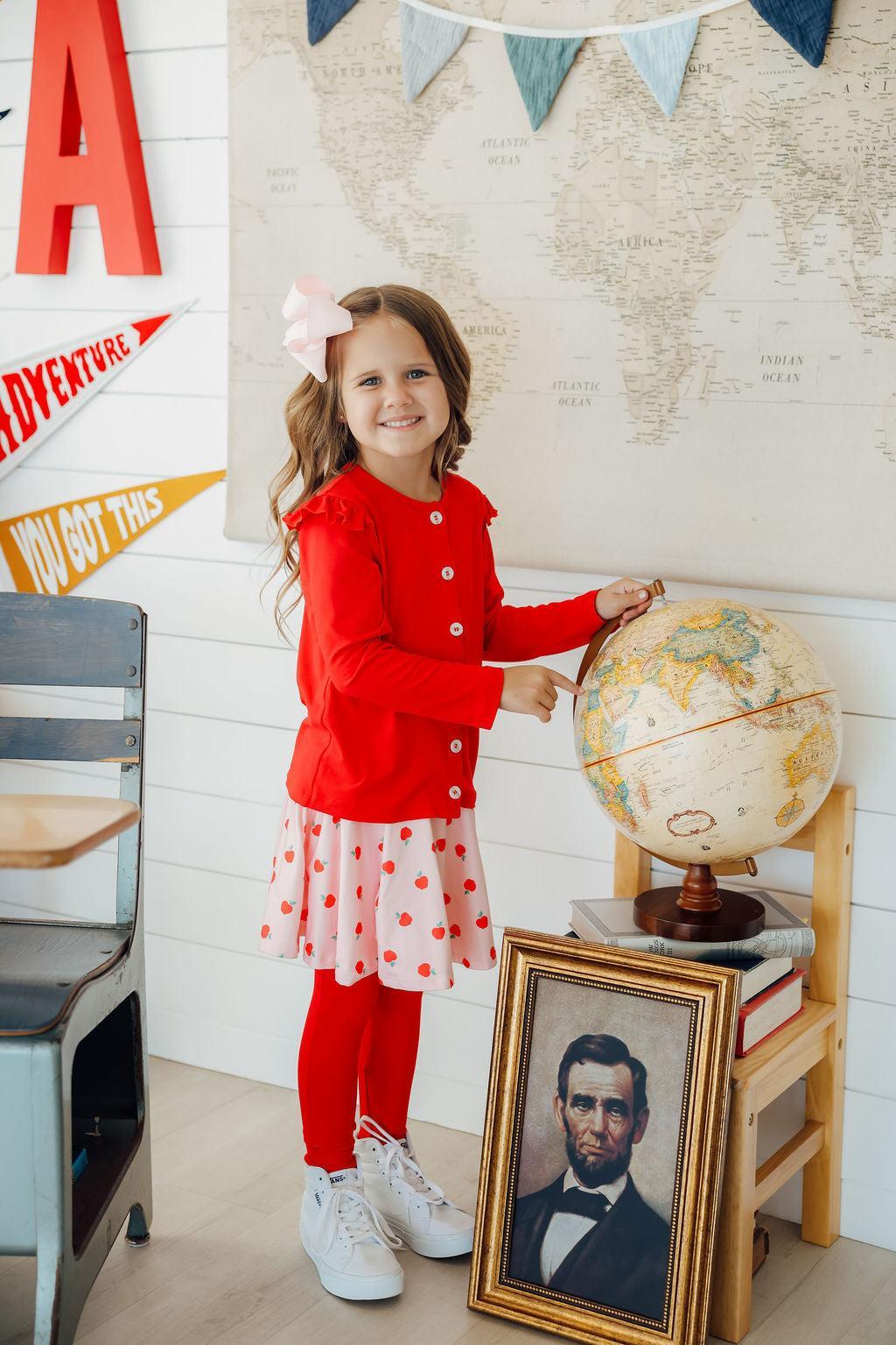 A smiling girl wearing An Apple a Day Twirl Skeggings for Girls stands by a globe, showcasing her vibrant outfit. These skeggings for girls combine comfort and style.