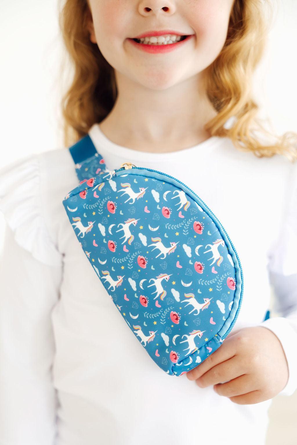 A young girl smiling while holding the Blue Moon Belt Bag, featuring a whimsical design with unicorns and colorful patterns on a charming blue background.
