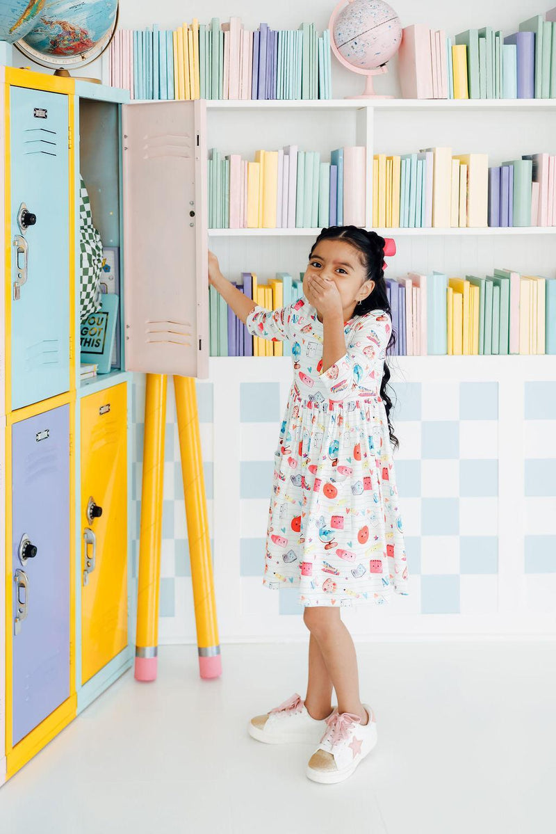 A cheerful girl in the Smarty Pants Pocket Twirl Dress from Mila & Rose, playfully covering her mouth, standing near pastel-colored lockers filled with books.