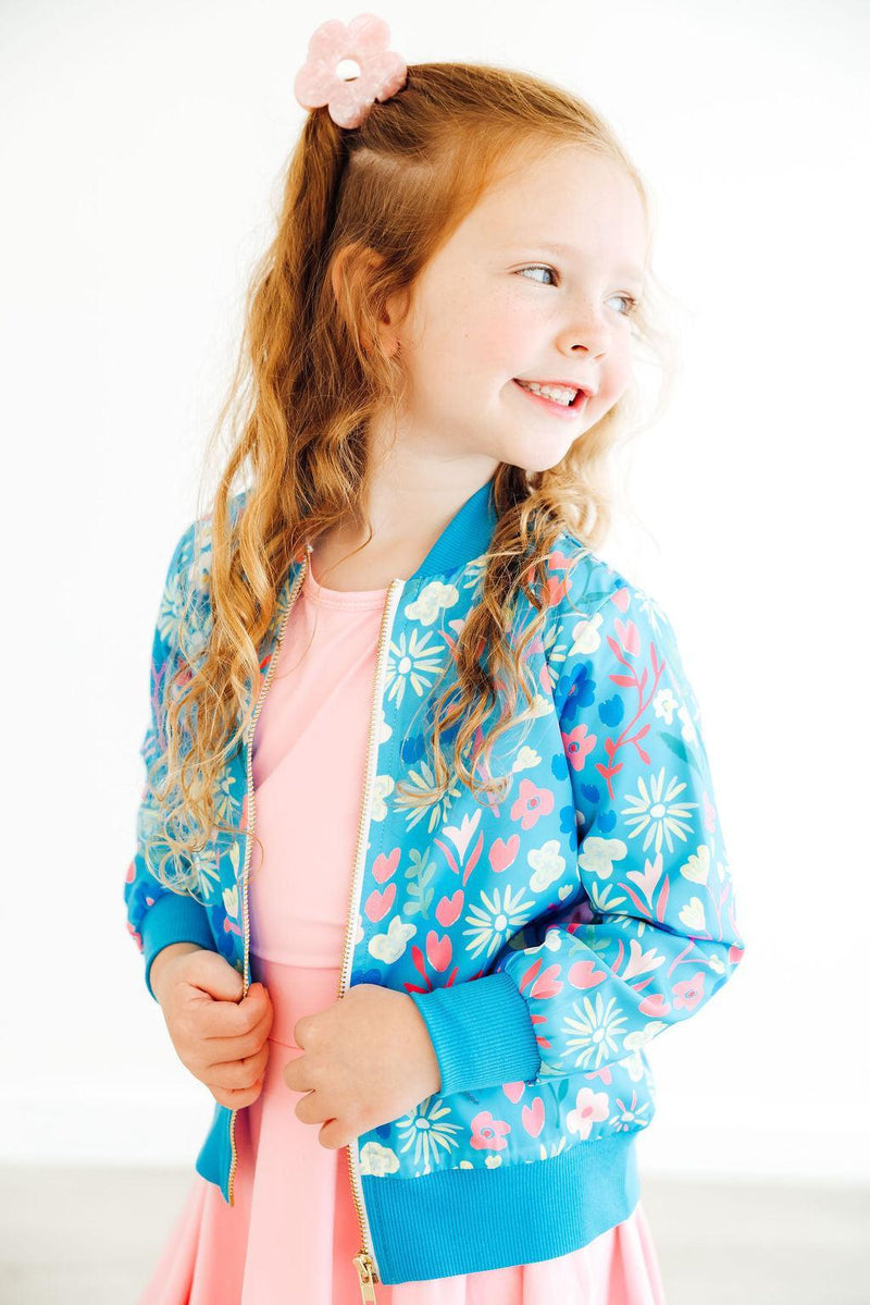A smiling girl wearing the Springtime Splendor Satin Jacket over a pink dress, expressing joy and charm in a bright, cheerful setting.