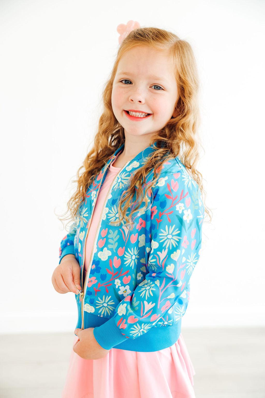 A cheerful girl wearing the Springtime Splendor Satin Jacket from Mila & Rose, featuring floral patterns, smiling while holding the jacket zipper, perfect for spring outfits.