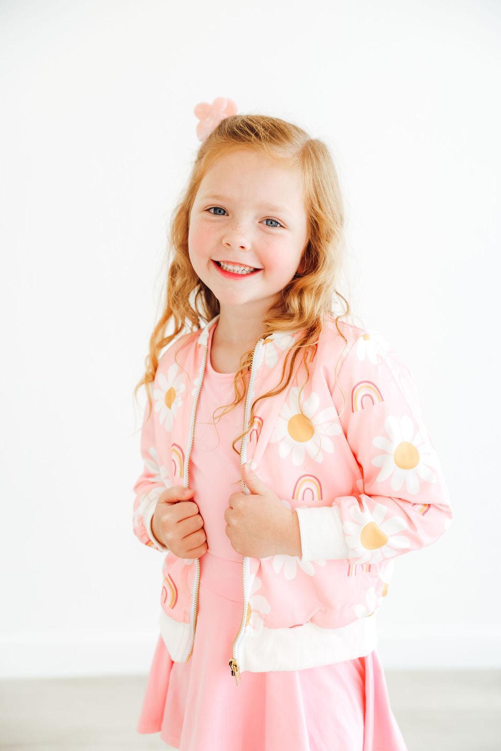 Girl wearing the You are My Sunshine Satin Jacket from Mila & Rose®, featuring a pink design with daisies and rainbows, smiling playfully in a bright, cheerful atmosphere.