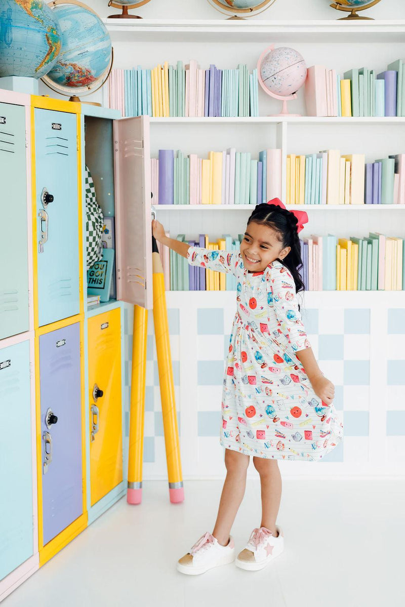 A happy girl wearing the Smarty Pants Pocket Twirl Dress from Mila & Rose, twirling playfully in front of colorful lockers, showcasing the dress's fun and vibrant design.