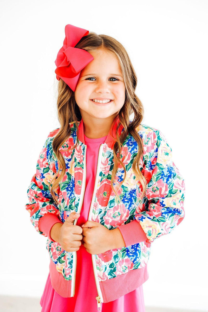 A cheerful girl in the Wildflower Wishes Satin Jacket by Mila & Rose, displaying its colorful floral design. She is smiling and posing with her hands on the jacket, radiating a fun and stylish vibe.
