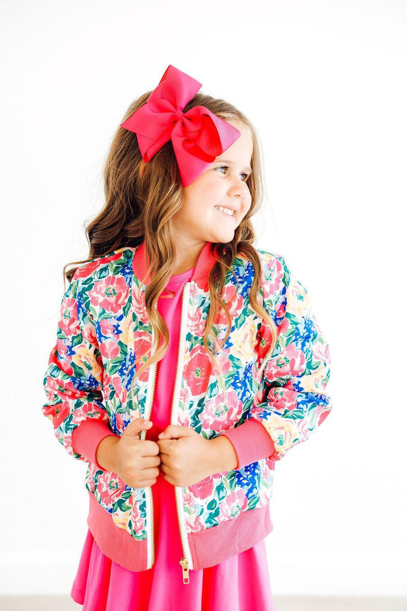 A young girl showcasing the Wildflower Wishes Satin Jacket from Mila & Rose with a vibrant floral pattern. She is looking to the side with a playful smile, adorned with a large pink bow in her hair.