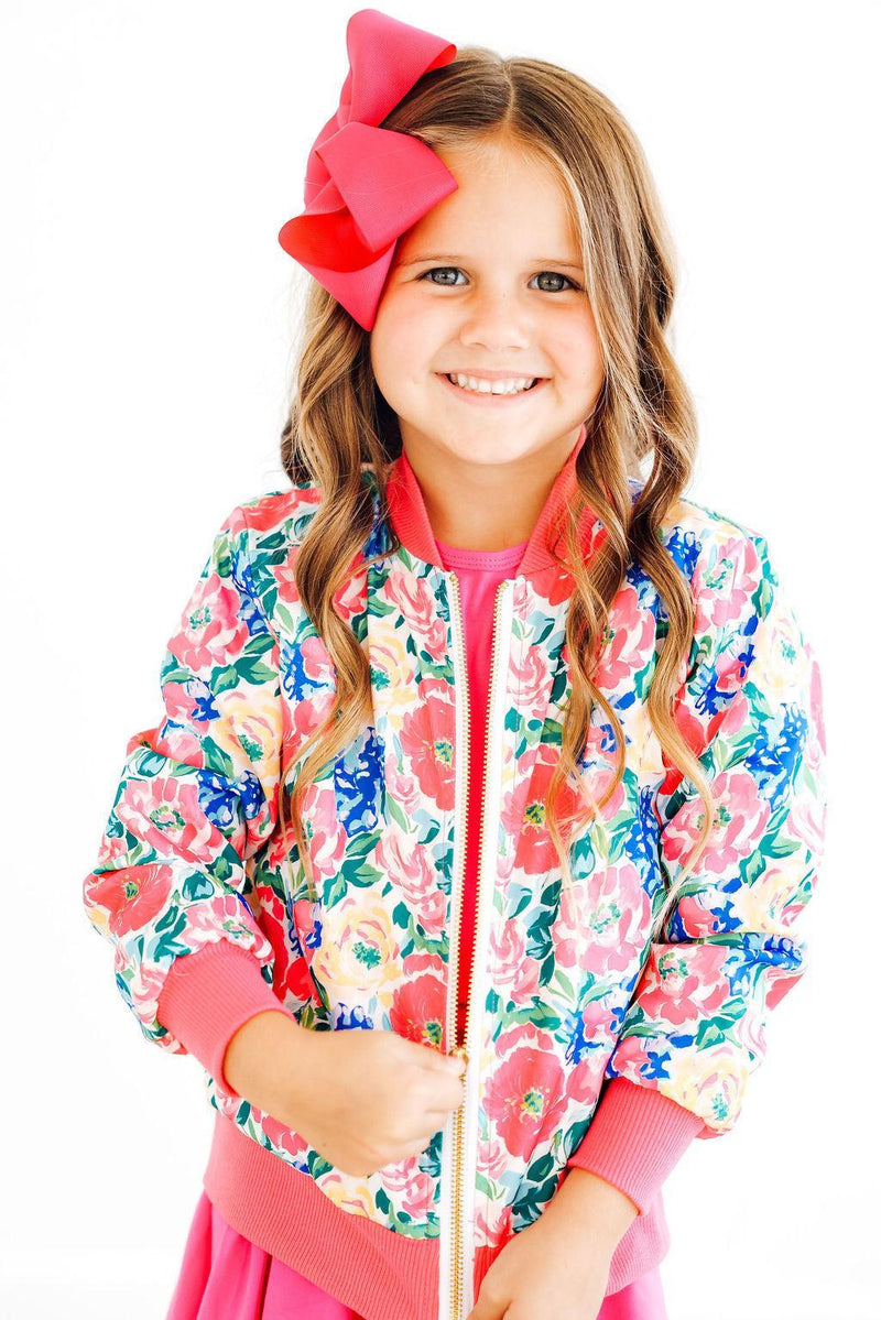 A young girl wearing the Wildflower Wishes Satin Jacket from Mila & Rose, featuring a colorful floral design. She is smiling brightly and holding the jacket with her hand, exuding joy and style.