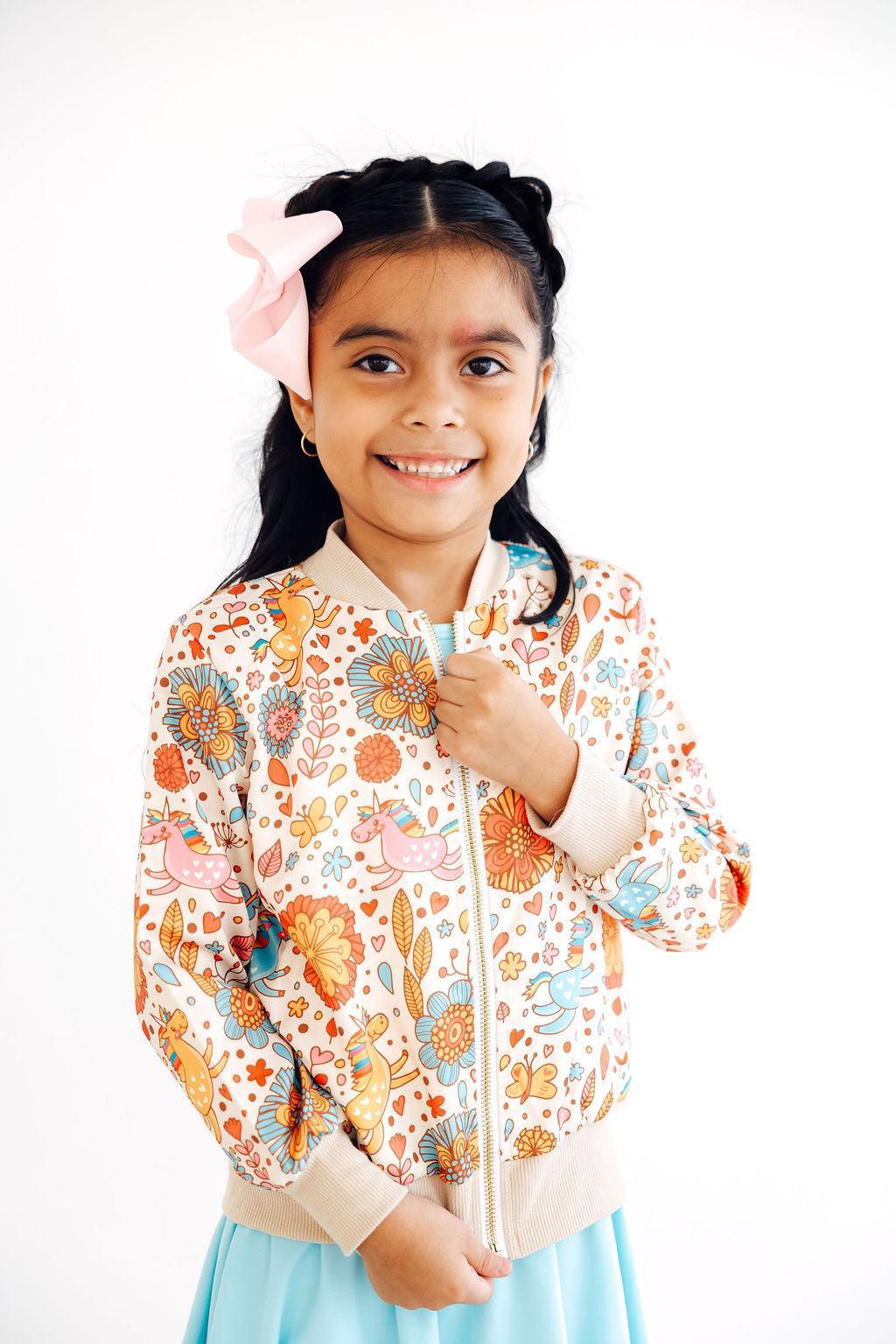 Smiling girl wearing the 'Retro Unicorns' satin jacket for girls, featuring a colorful floral design, styled with a twirl dress.