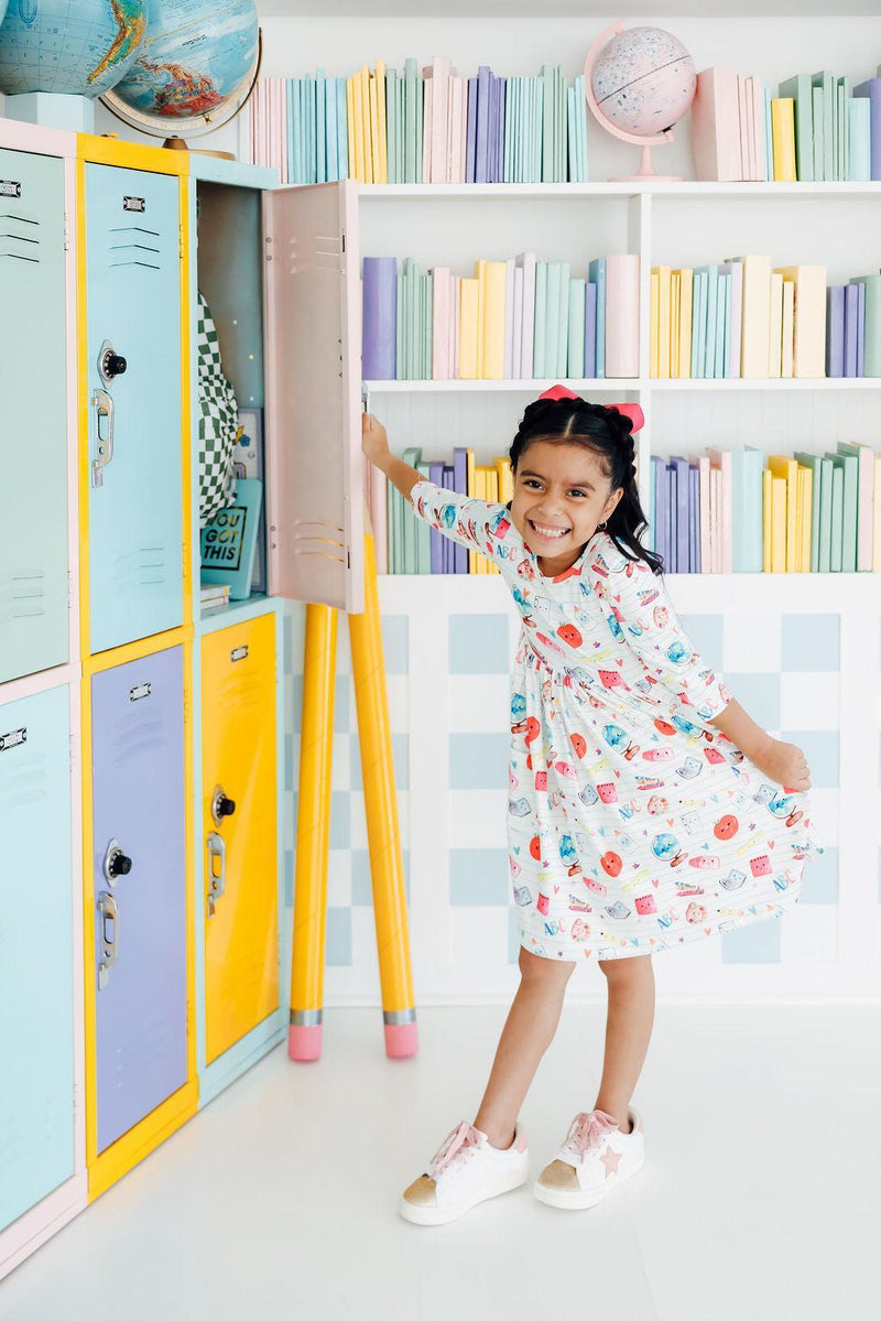 A joyful girl in the Smarty Pants Pocket Twirl Dress by Mila & Rose, posing with a big smile next to a vibrant locker set, showcasing her playful outfit.