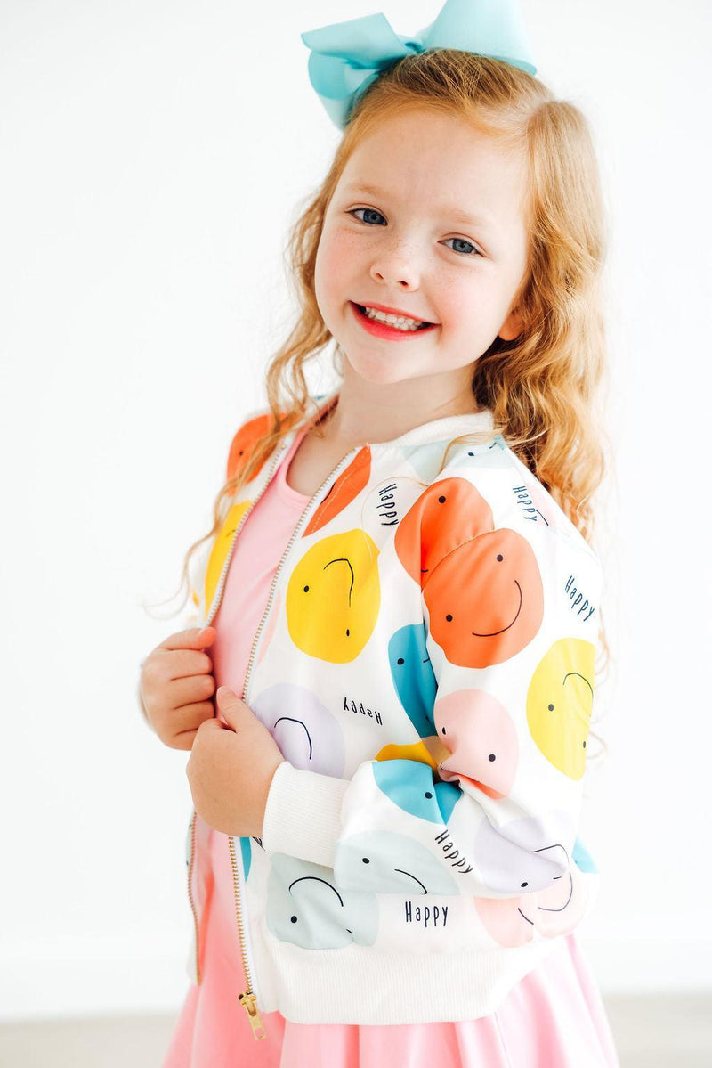 A young girl happily posing in the 'Happy Days' Satin Jacket for Girls, featuring a mix of colorful smiley faces, complemented by her pink dress.