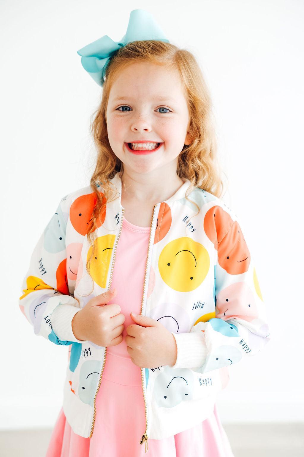 A smiling girl wearing the 'Happy Days' Satin Jacket for Girls, featuring colorful smiley faces, paired with a pink dress, showcasing a cheerful and playful look.