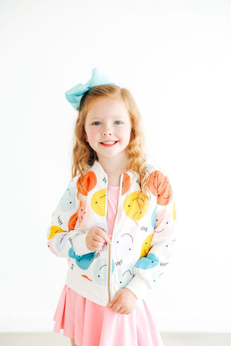 Cheerful girl wearing the 'Happy Days' Satin Jacket for Girls with smiley faces, zipping up her jacket while looking joyful in a pink dress.