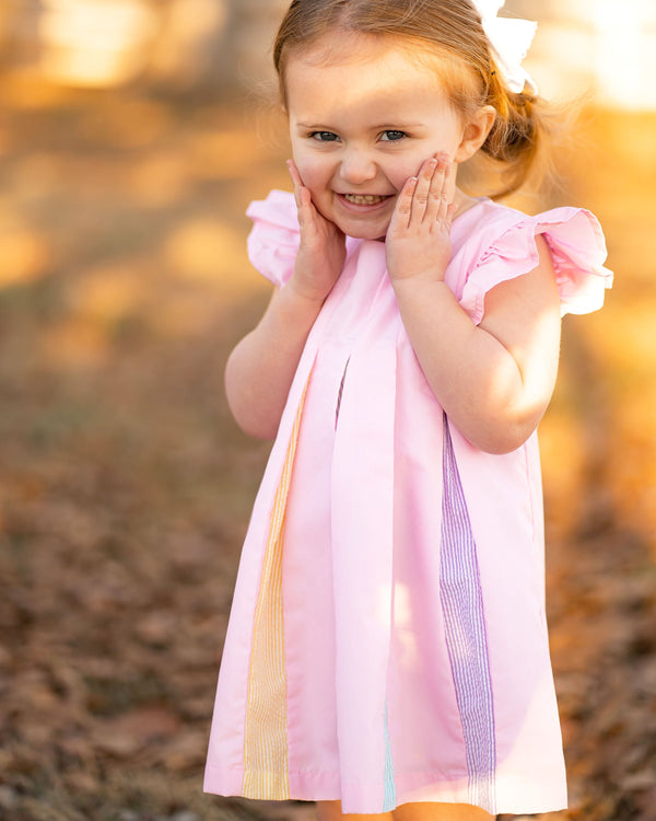 Smiling girl in pink swing dress with flutter sleeves and colorful rainbow pleat accents down the front.