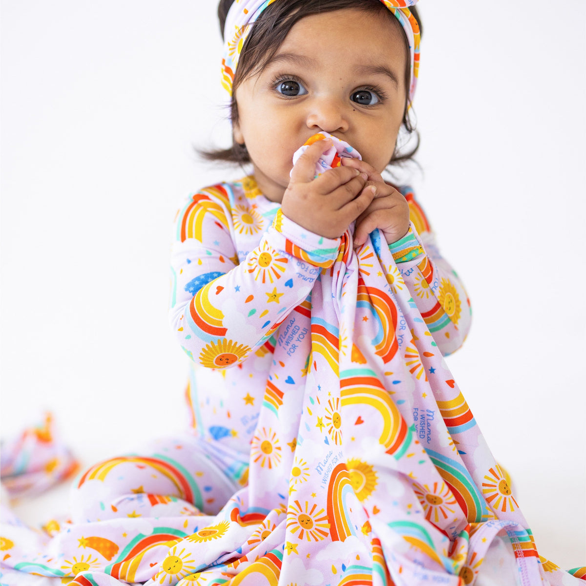 A joyful baby biting on The Forever Swaddle Blanket | Here Comes the Sunshine (Pink), showcasing colorful rainbows and sun motifs.