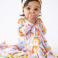 A joyful baby biting on The Forever Swaddle Blanket | Here Comes the Sunshine (Pink), showcasing colorful rainbows and sun motifs.