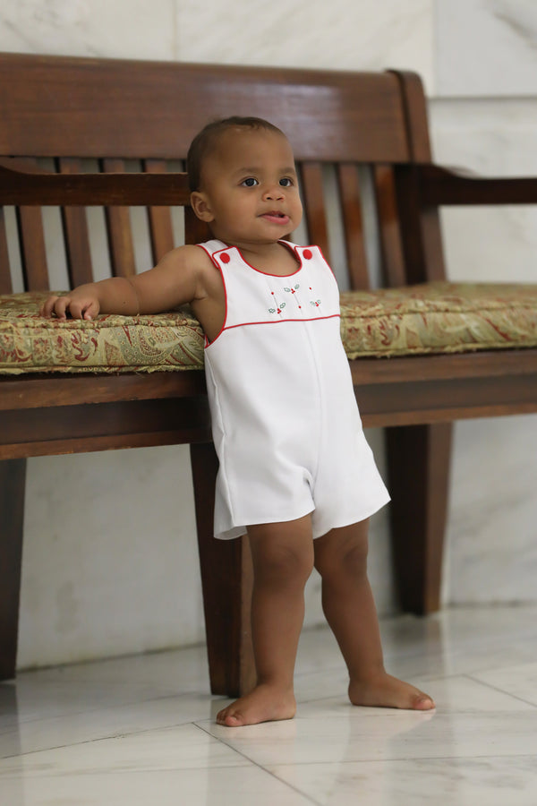 Toddler wearing white holiday romper with embroidered holly, red trim, and pleated yoke standing by wooden bench.