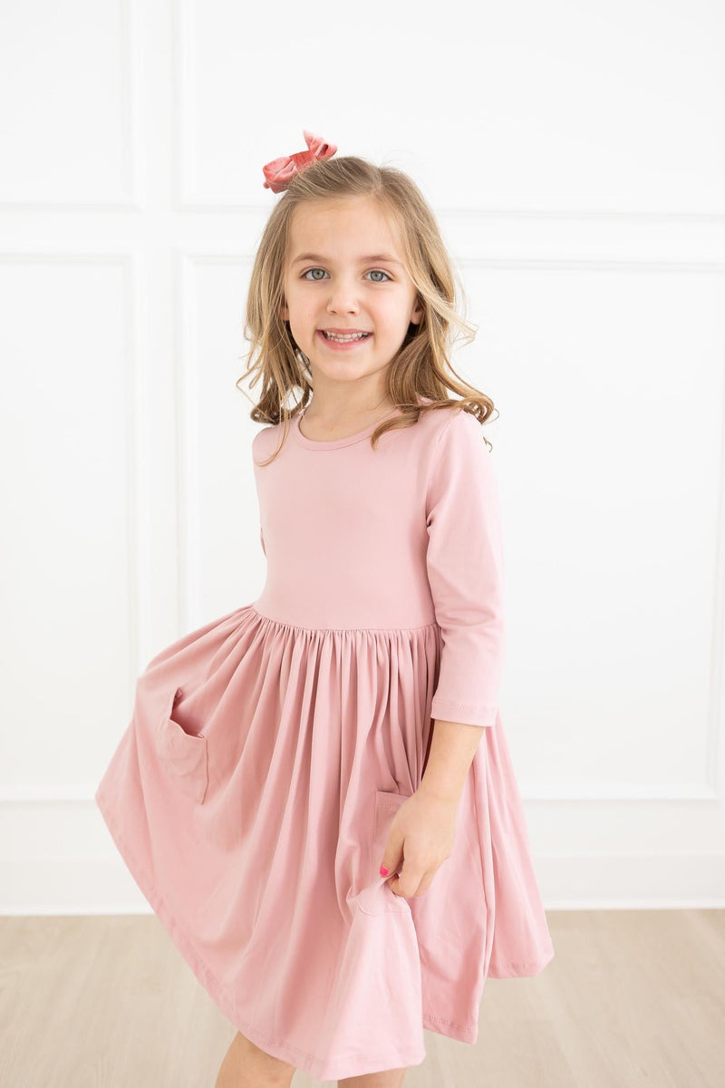 A smiling young girl wearing a Vintage Pink 3/4 Sleeve Pocket Twirl Dress by Mila & Rose®, showcasing its playful design and soft fabric, perfect for twirling and play.
