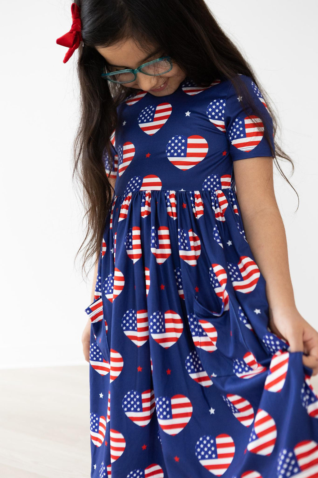 A girl wearing the 'I Heart the USA' Short Sleeve Pocket Twirl Dress for Girls, showcasing a cheerful print of hearts with American flags, perfect for twirling.