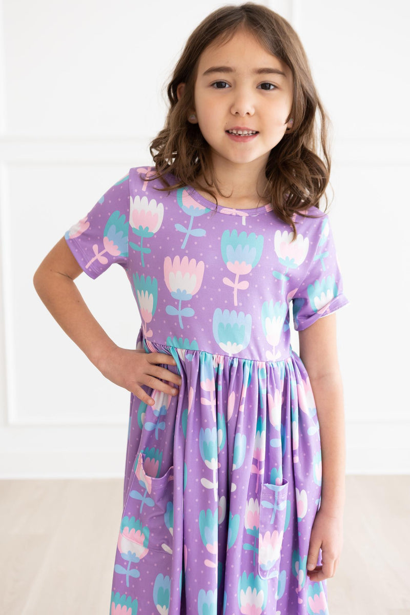 Girl modeling the Too Cool Tulips S/S Pocket Twirl Dress, showcasing a vibrant tulip print with pockets, perfect for playful occasions.