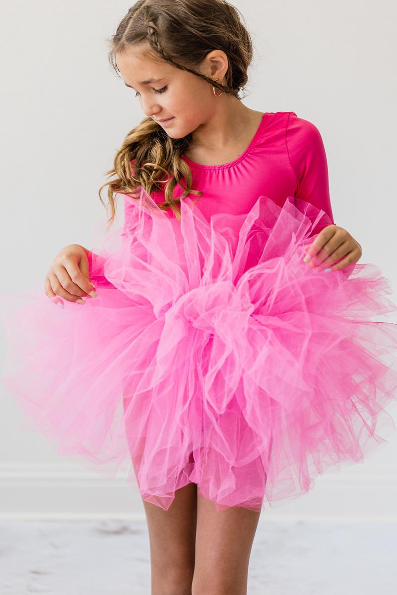 A young girl inspecting her Hot Pink 3/4 Sleeve Fluffy Tutu Leotard for Girls, highlighting the fluffy tulle skirt. This tutu leotard for girls adds a cheerful touch to any dance outfit.