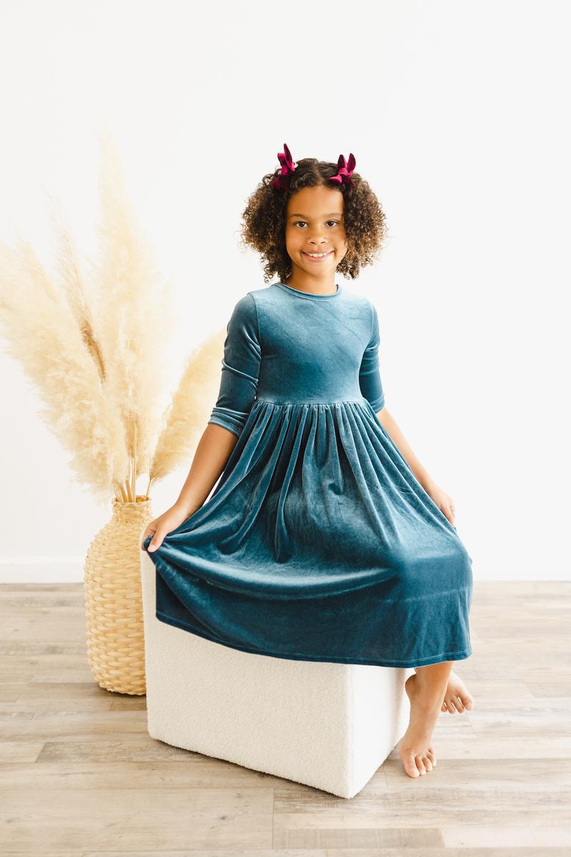 Girl seated on a cube, wearing the Denim Blue Velvet Twirl Dress for Girls, showcasing the elegant style and cozy design in a bright setting.
