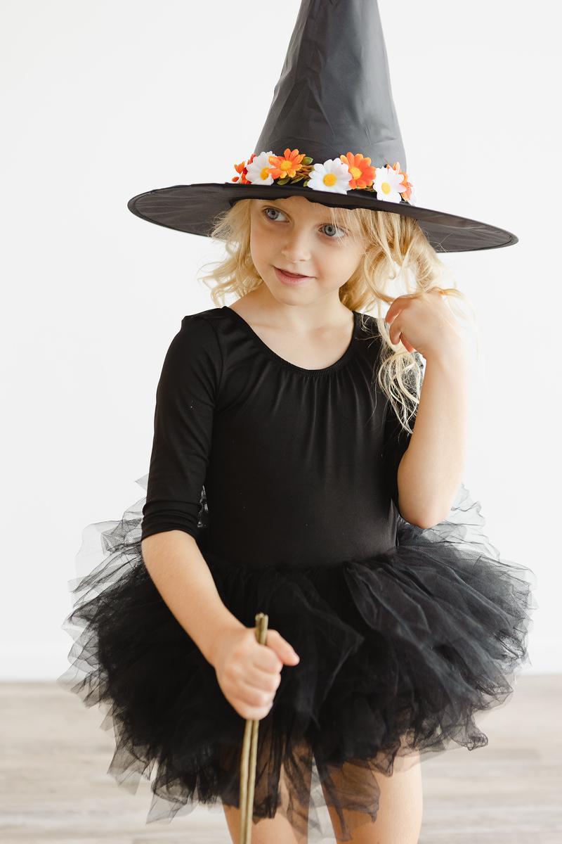 A girl in a Black 3/4 Tutu Leotard by Mila & Rose, looking charming with a witch hat and playful smile, embodying the joyful spirit of dance.