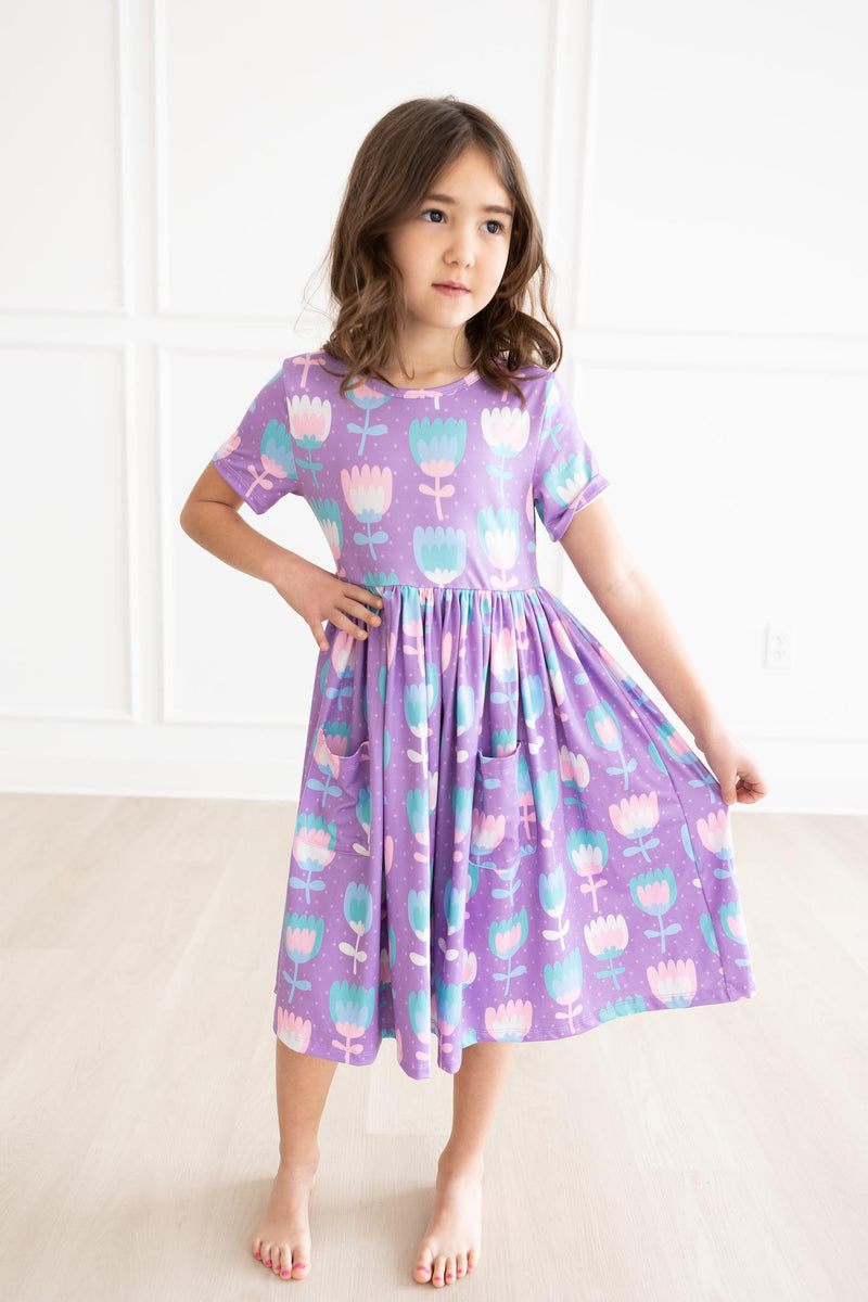 Girl in a playful pose wearing the Too Cool Tulips S/S Pocket Twirl Dress, featuring a cheerful floral pattern, perfect for twirling and fun activities.