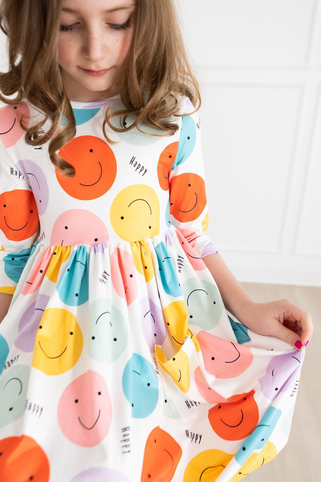 A young girl wearing the 'Happy Days' 3/4 Pocket Fun Twirl Dress for Girls, featuring colorful smiley faces, showcasing the playful design and twirl effect.