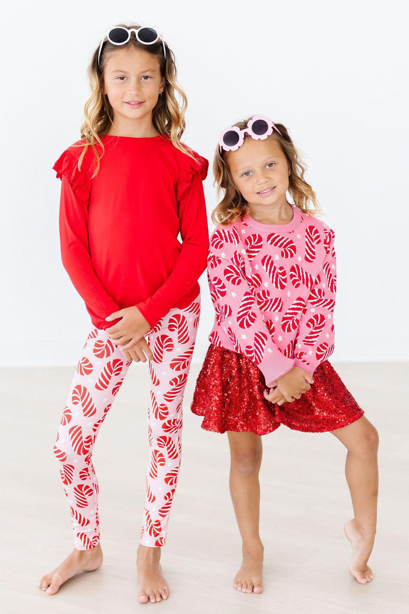 Two smiling girls wearing festive outfits, showcasing the Candy Canes Sweater from Mila & Rose. One girl is in a red top with candy cane leggings, and the other in a pink sweater with a sparkly red skirt, perfect for holiday celebrations.