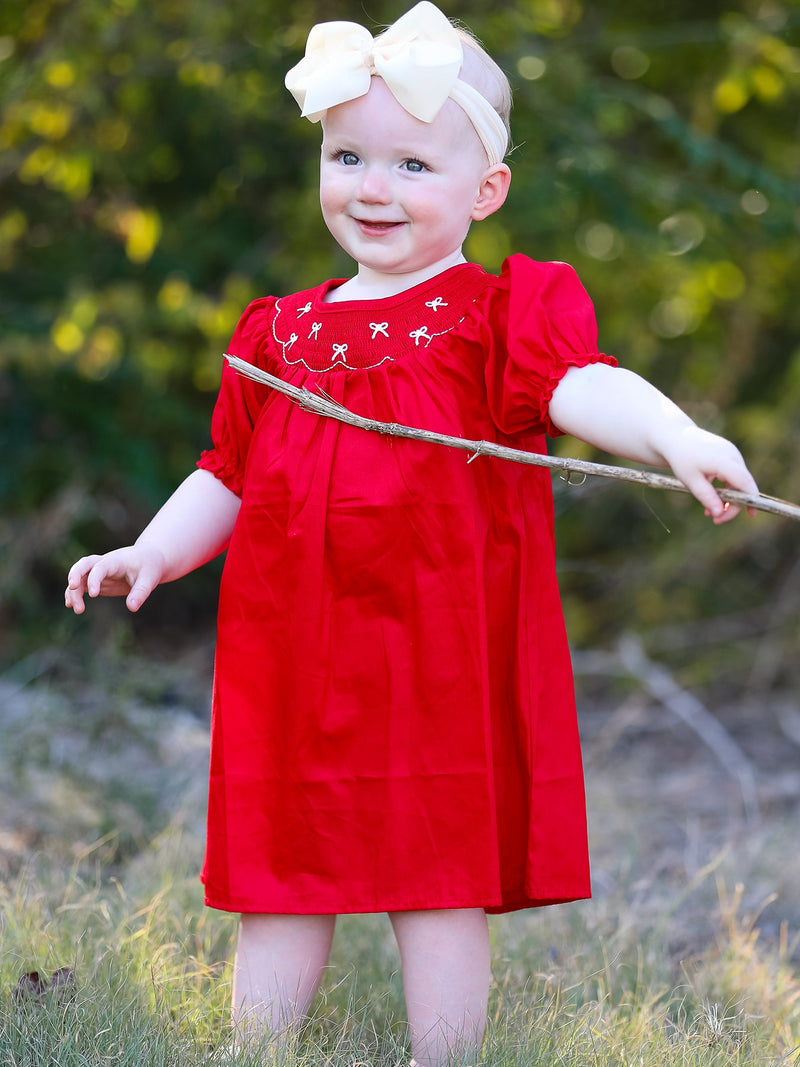 Smiling toddler in a red bow smocked embroidered dress with a cream headband bow outdoors.