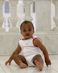 Baby boy in white cotton pique holiday romper with red trim, pleated yoke, and embroidered holly details.
