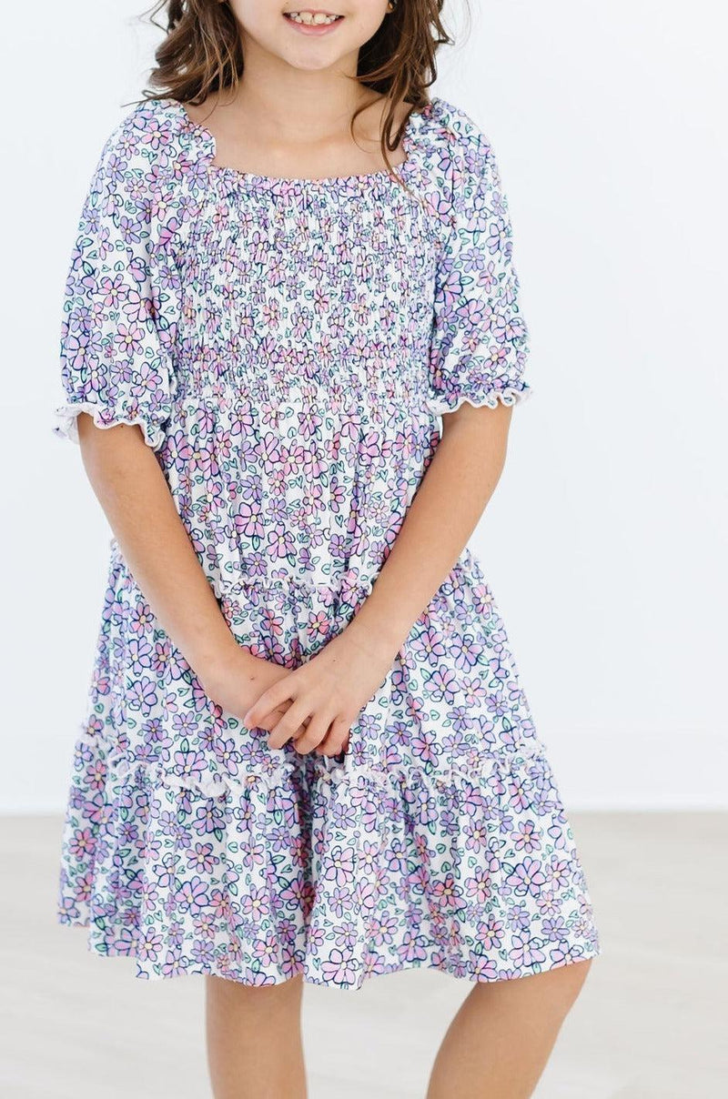 A girl wearing the Lovely Lavender Smocked Ruffle Dress, radiating joy with a charming smile and stylish attire.