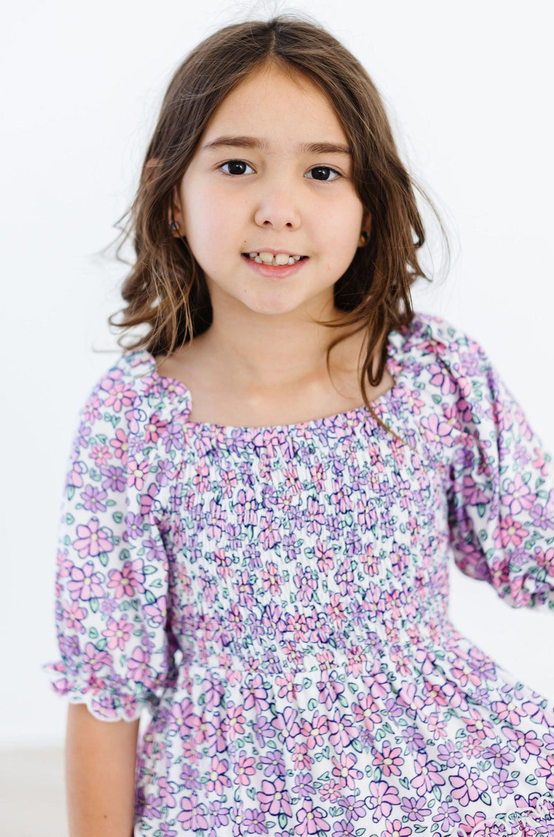 Close-up of a smiling girl wearing the Lovely Lavender Smocked Ruffle Dress, highlighting the charming smocked bodice and vibrant floral details.