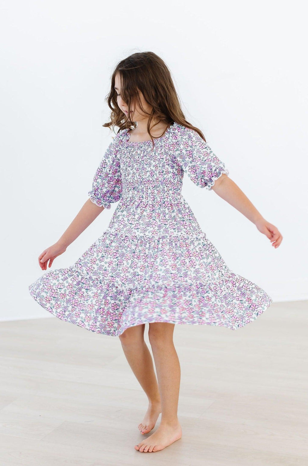 A joyful child twirling in the Lovely Lavender Smocked Ruffle Dress, showcasing its charming floral pattern and playful ruffles, perfect for spring adventures.