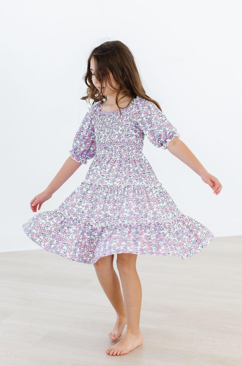 A joyful child twirling in the Lovely Lavender Smocked Ruffle Dress, showcasing its charming floral pattern and playful ruffles, perfect for spring adventures.