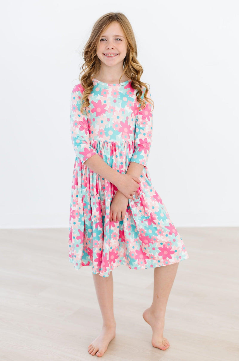 A smiling girl wearing the Petal Perfection 3/4 Sleeve Pocket Twirl Dress, highlighting its colorful floral pattern and twirl-worthy design, perfect for fun and playful moments.