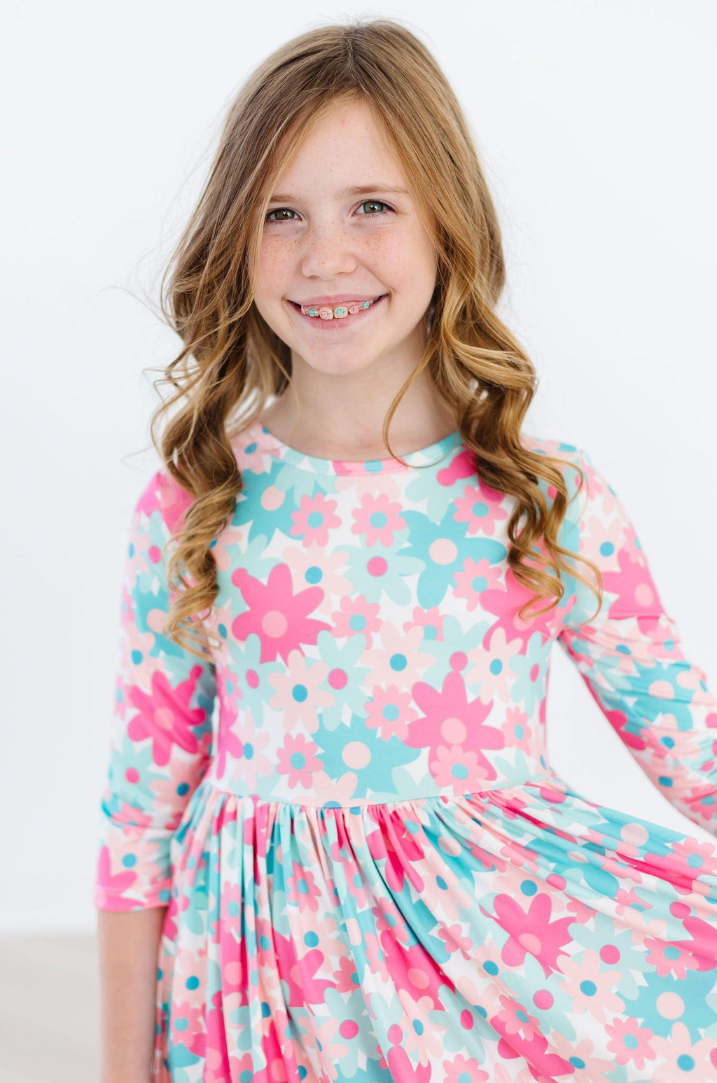 A cheerful girl wearing the Petal Perfection 3/4 Sleeve Pocket Twirl Dress, featuring a vibrant floral pattern in pink and blue, showcasing her joyful smile and playful spirit.