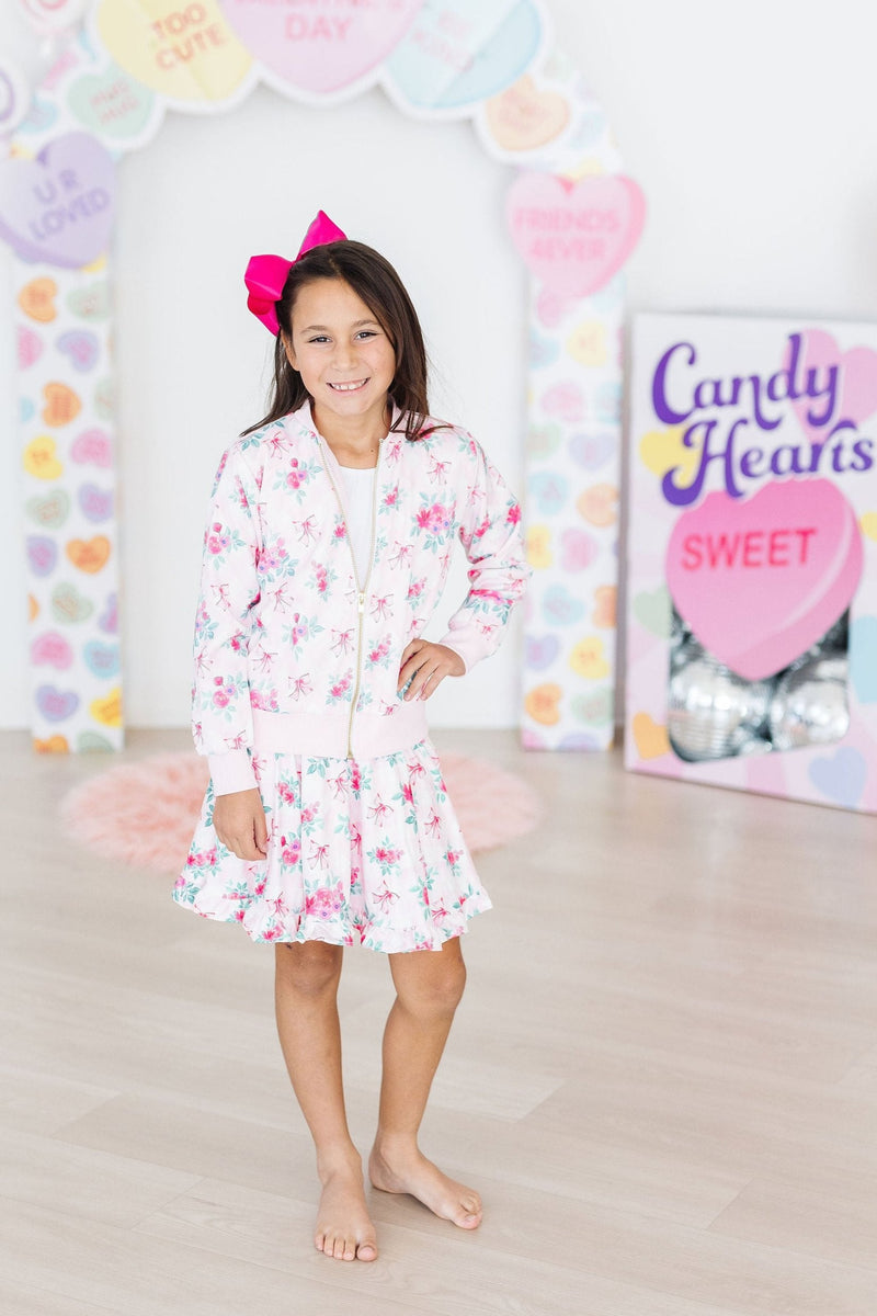 A cheerful child wearing the Gingham Bows Ruffle Twirl Skort by Mila & Rose, posing with a pink floral bomber jacket, surrounded by a colorful heart-shaped décor.