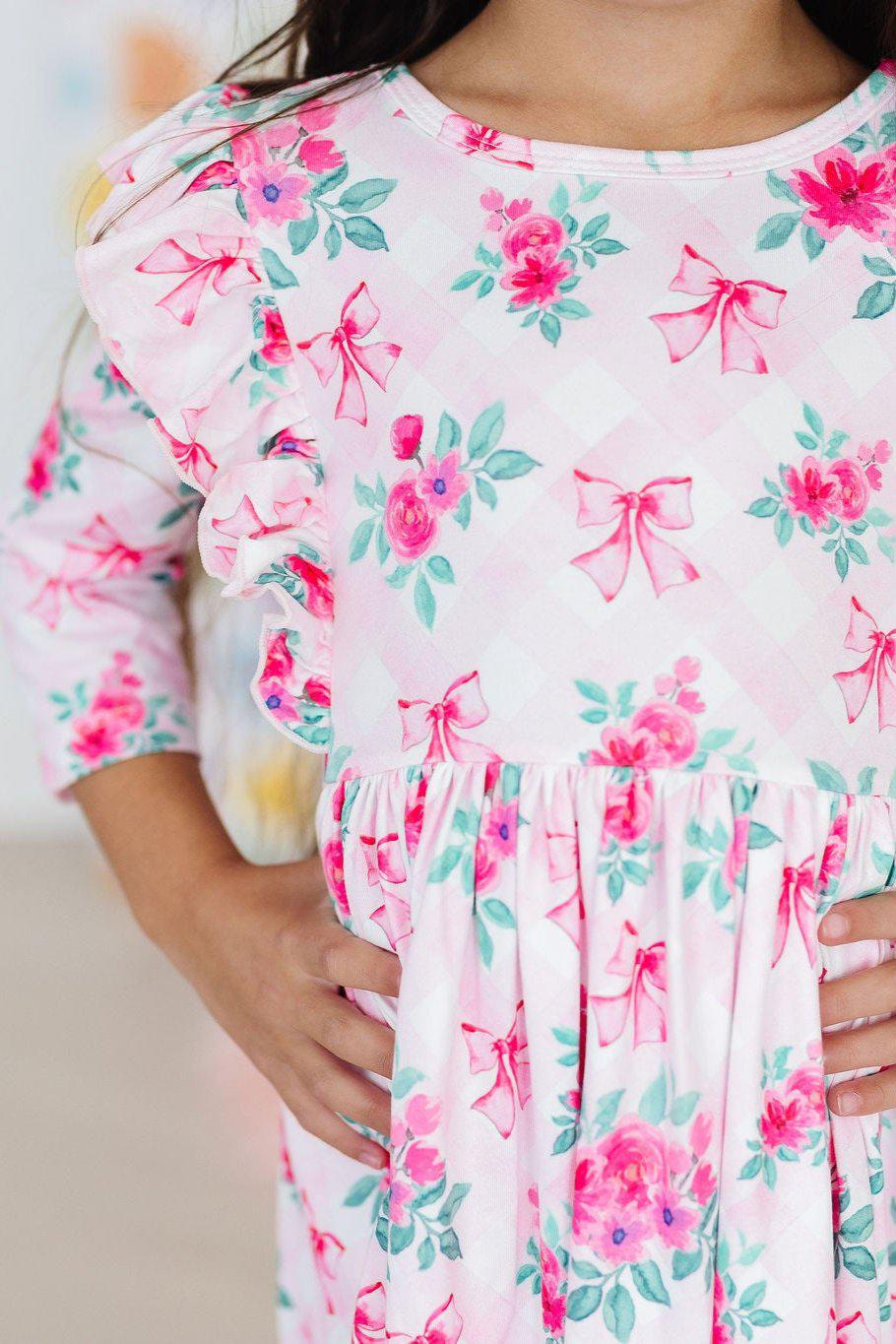 Adorable girl wearing the Gingham Bows 3/4 Sleeve Ruffle Twirl Dress with floral prints, showcasing the dress's playful ruffles and twirlability, perfect for every little fashionista.