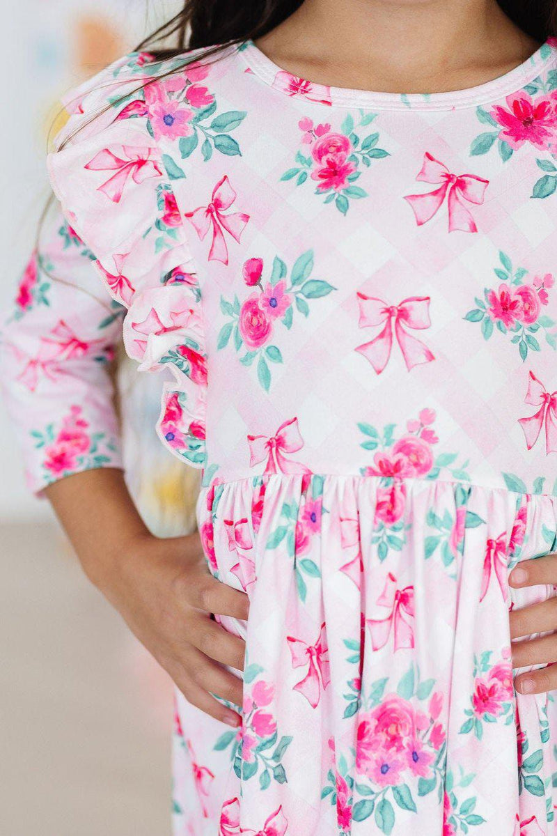 Adorable girl wearing the Gingham Bows 3/4 Sleeve Ruffle Twirl Dress with floral prints, showcasing the dress's playful ruffles and twirlability, perfect for every little fashionista.